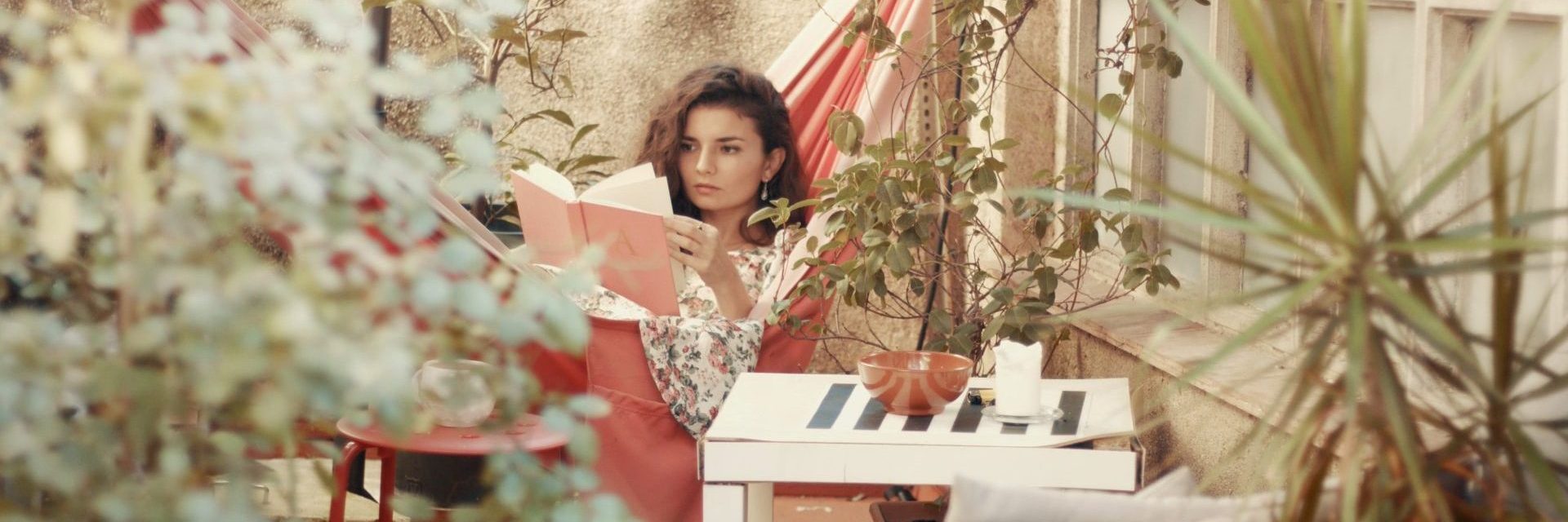 Eine Frau sitzt auf einem von Pflanzen umgebenen Balkon und liest ein Buch. Im Hintergrund sind ein Sonnenschirm und Gartenmöbel zu sehen.