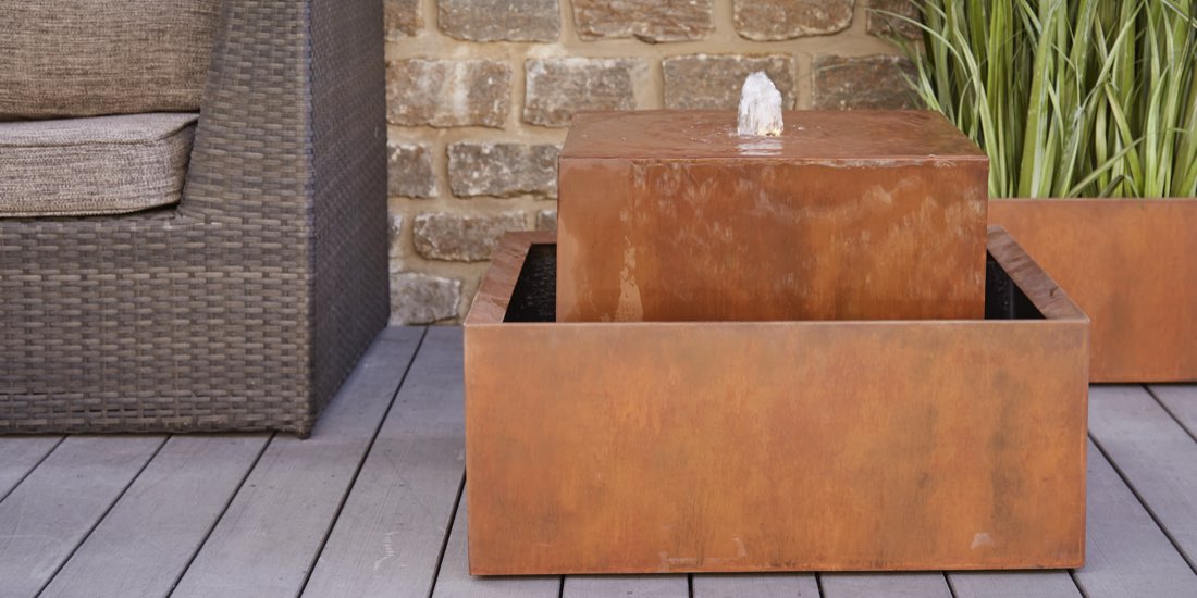 Ein moderner Gartenbrunnen aus Cortenstahl: Er ist rechteckig, rostbraun und das Wasser quillt oben aus der Mitte des Blocks. Er steht auf einer Terrasse im Außenbereich.