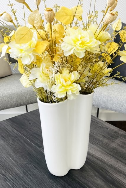 Ein Strauß vornehmlich gelber Kunstblumen in einer cremefarbenen Blumenvase, die eine organisch geschwungen Form besitzt